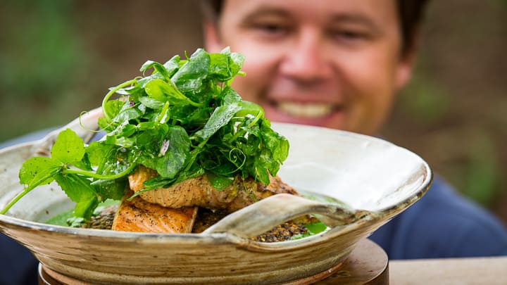 Eric Wilson smiles as he looks at two Grilled Lemon Salmon fillets stacked on top of Wilted Spicy Greens and Crispy Wild Rice Pancakes. A vibrant green Garden Puree surrounds the outside in the serving platter and the dish is topped with a pea tendril salad.