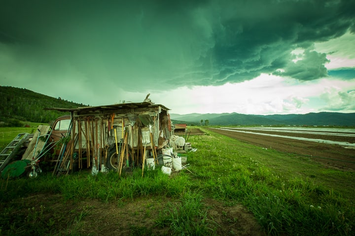 An old farm truck is converted into a tool shed and parked by the edge of farm rows. The wide vista across the farm to the mountains beyond is darkened by a rain storm and clouds above.