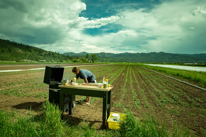 A butcher block table and grill are set up in the field of a farm. Rows of early season crops stretch into the distance and a backdrop of mountains and a cloud filled sky