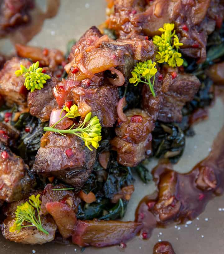 Bite sized crispy pork belly pieces sit on a bed of pork infused collard greens, a spicy dark red rhubarb compote is drizzled on top and bright yellow mustard flowers add a pop of color to the plated dish. 
