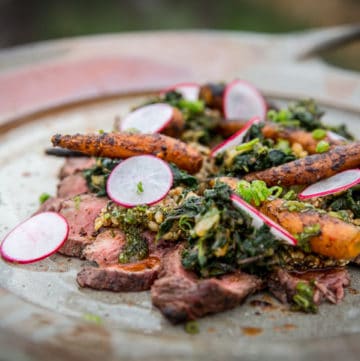 Looking down on a hand made platter with slices of a grilled petite tender covered with a scattering of grilled baby carrots, vibrant radish slices and piles of dark green Kimchi