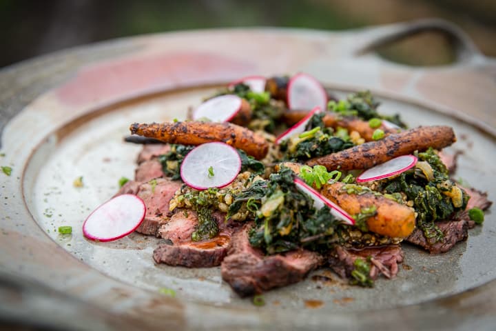 Looking down on a hand made platter with slices of a grilled petite tender covered with a scattering of grilled baby carrots, vibrant radish slices and piles of dark green Kimchi