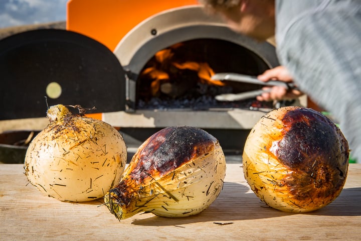 Three Hay-Roasted Onions fresh out of the oven sitting on cutting board in front of wood-fired oven in background