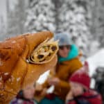 A gloved hand holds up a Backcountry Curry Chicken Salad wrap in front of a wintery family picnic scene