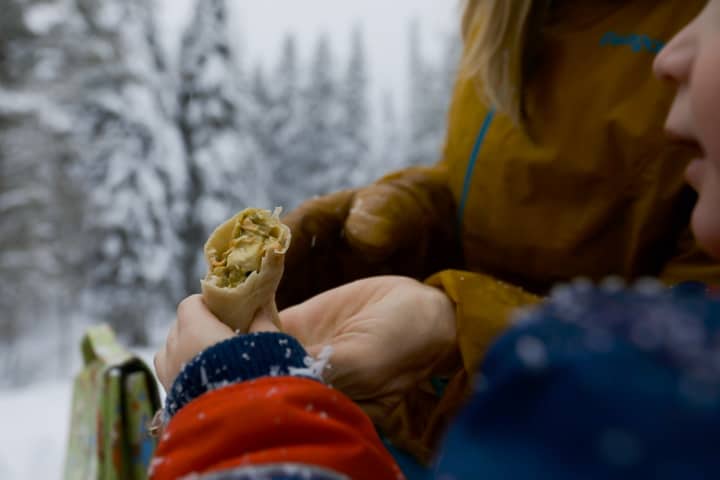 A kids hand holds up a Backcountry Curry Chicken Salad wrap during a wintery family picnic scene