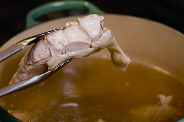 A pair of tongs hold a poached chicken breast over a pot of chicken stock for the Backcountry Curry Chicken Salad.